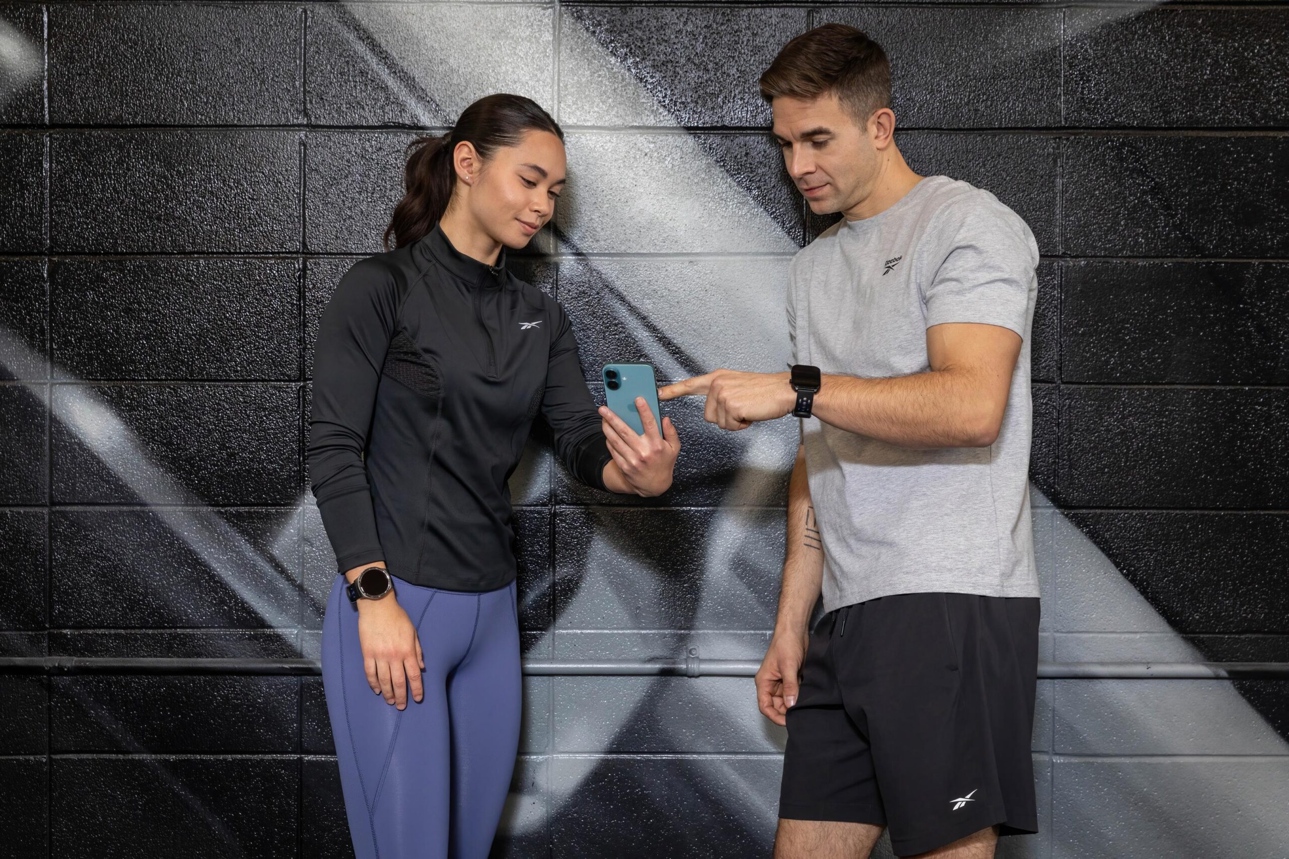 “Two people using a smartphone and wearing Reebok smartwatches at the gym.”